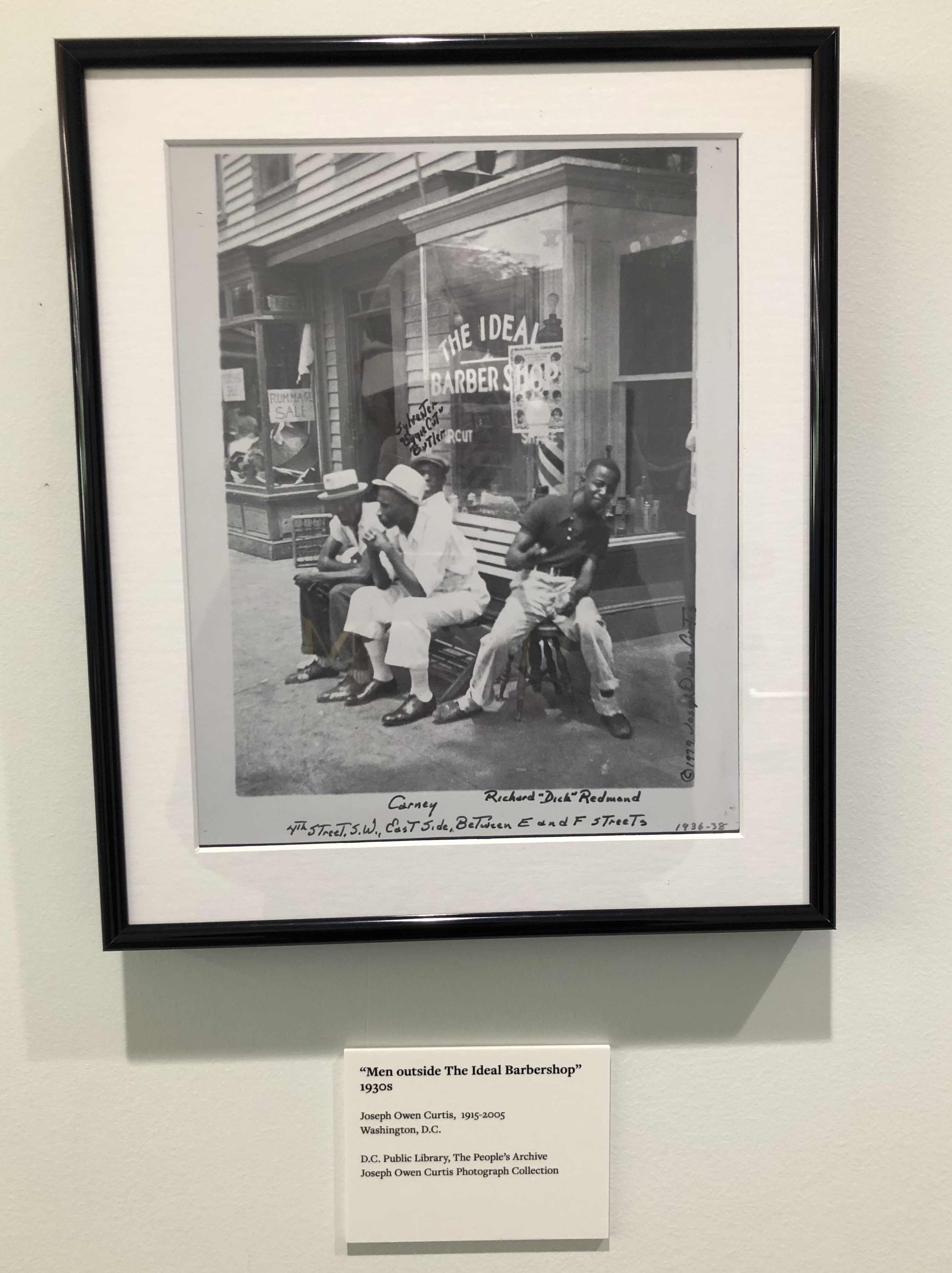 Photo of a framed 1930’s black-and-white photograph hung on a white wall. The framed photo depicts four African American men of varying ages sitting on a bench outside of a glass store windowfront which reads “The Ideal Barbershop.” The location is written on the photo in black ink.