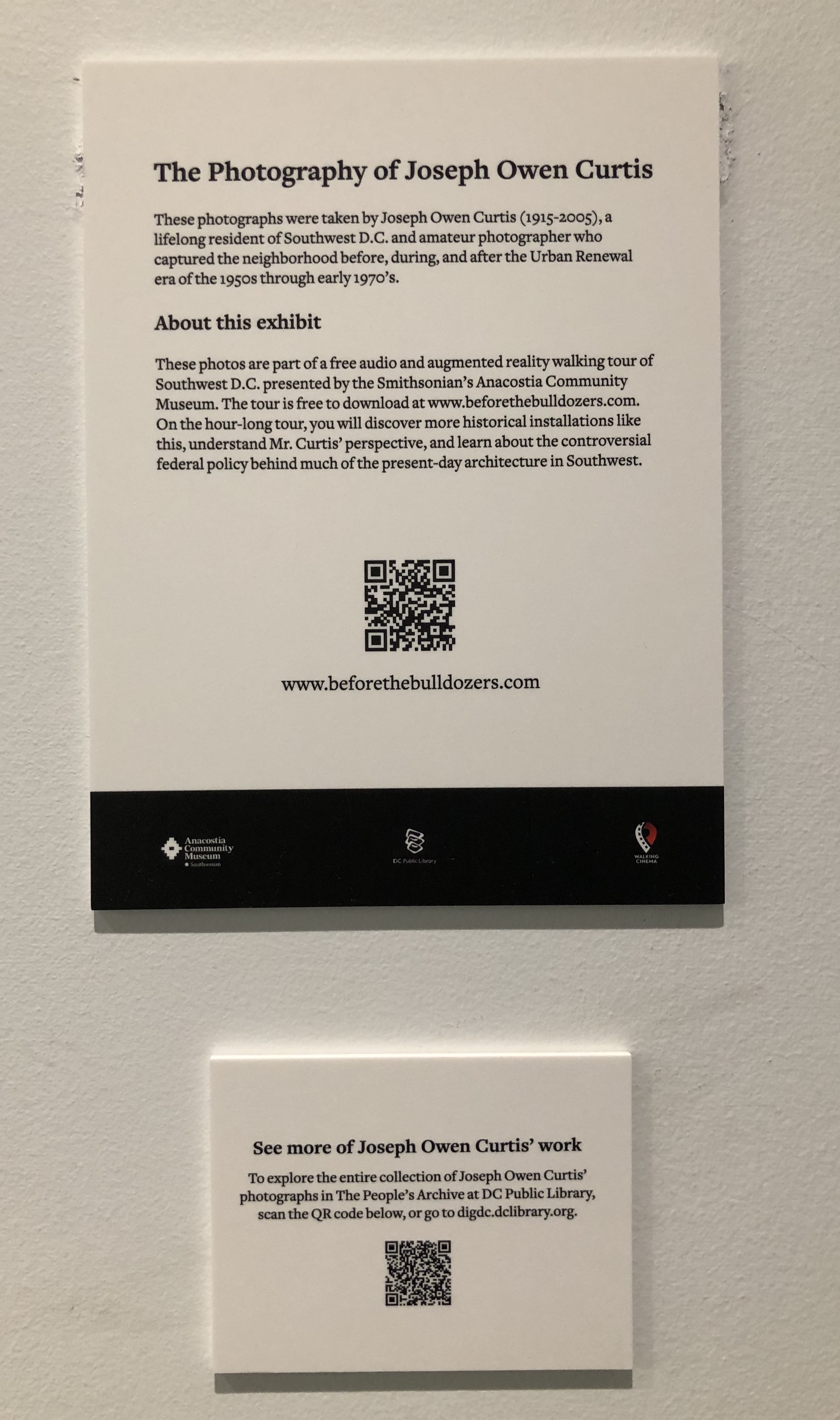 Photograph of two white wall plaques. The larger plaque has a heading that reads “Photography of Joseph Owen Curtis,” and a square QR code. The smaller plaque reads “See more of Joseph Owen Curtis’s work” and has another small QR code.