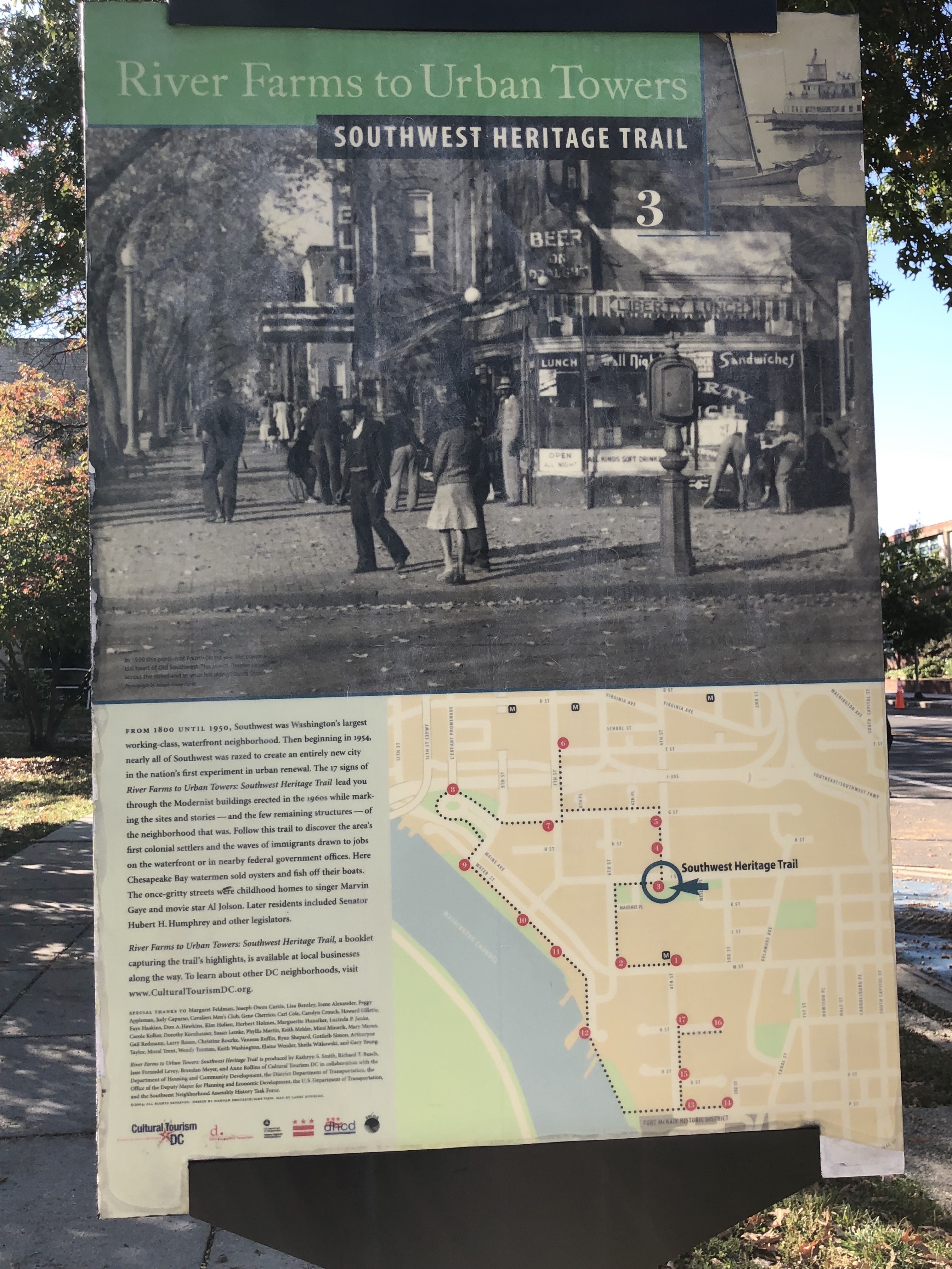 Photograph of a large sidewalk sign with the heading “River Farms to Urban Towers: Southwest Heritage Trail.” A large black-and-white photograph of a sidewalk with shops and trees takes up the top-half of the sign, with small text and an illustrated map on the bottom half.
