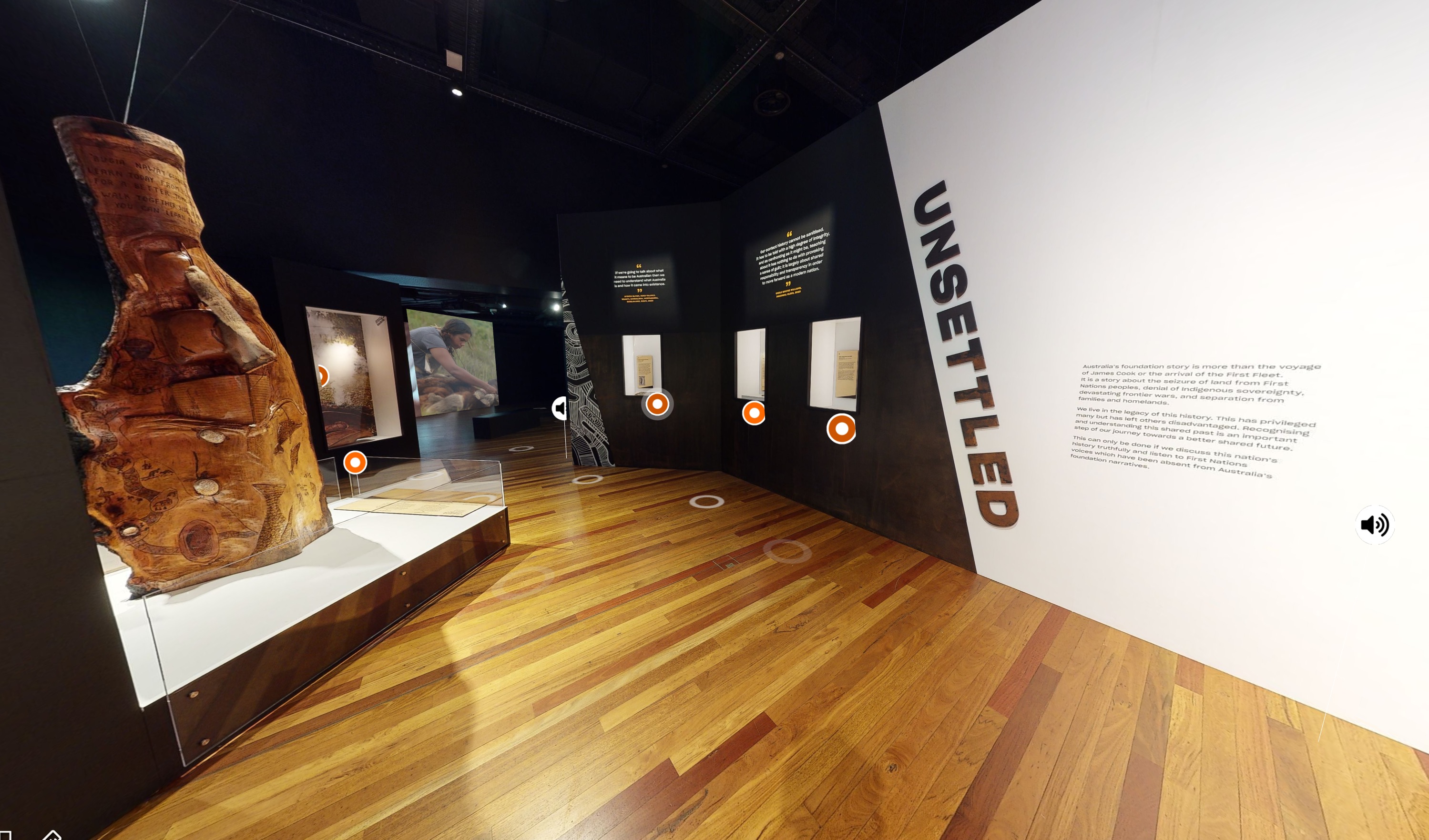 A screenshot taken from the virtual tour of the Australian Museum’s Unsettled exhibit. The gallery space has wooden floors and black and white walls. Displayed on these walls are various objects and text for object labels. Below each of these objects is an orange circle for visitors to click on for more information.