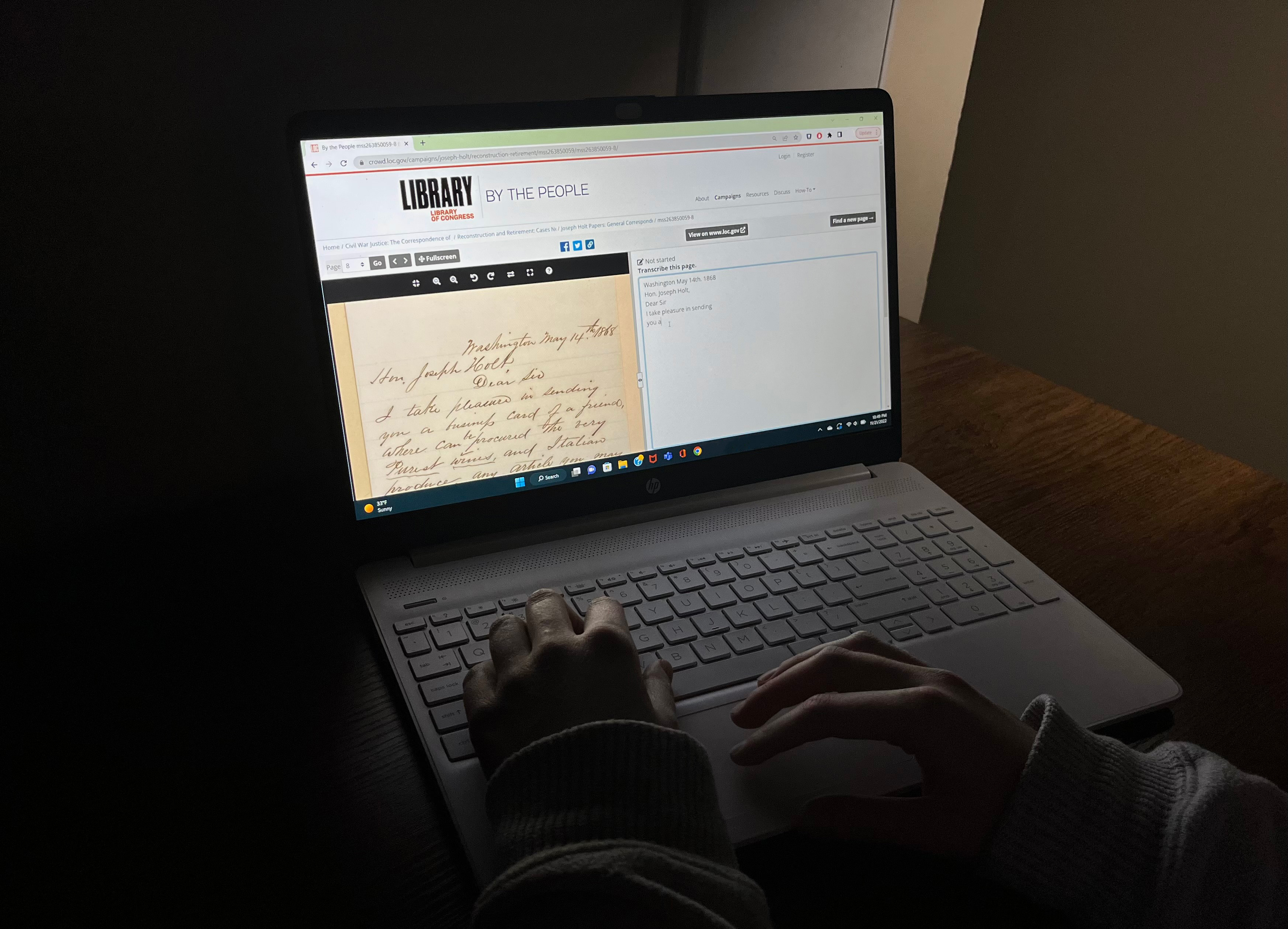 In a dark room, hands type on the keyboard of a laptop displaying a webpage with the text “Library of Congress” “By the People.” The webpage has a historic document next to a transcription box with text in it.
