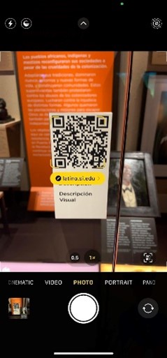 Screenshot of an iPhone camera view that shows a yellow pop-up link that reads “latino.si.edu” emerging from a QR code on an exhibit display with the label “visual description” underneath.