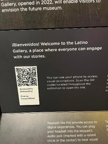 Black and white QR code with the label “Accessibility Onboarding” posted on a black panel with instructive white text. The top of the panel reads “¡Bienvenidos! Welcome to the Latino Gallery, a place where everyone can engage with our stories.” The text to the write of the code reads “You can use your phone to access visual descriptions. Scan the QR codes located throughout the exhibition to open the link.”