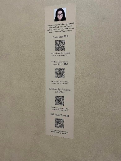Semi-blurry image of a light-brown vertical text panel on a darker beige wall. On the panel is an image of a woman, some text, and four QR codes stacked vertically with various tour names above each.