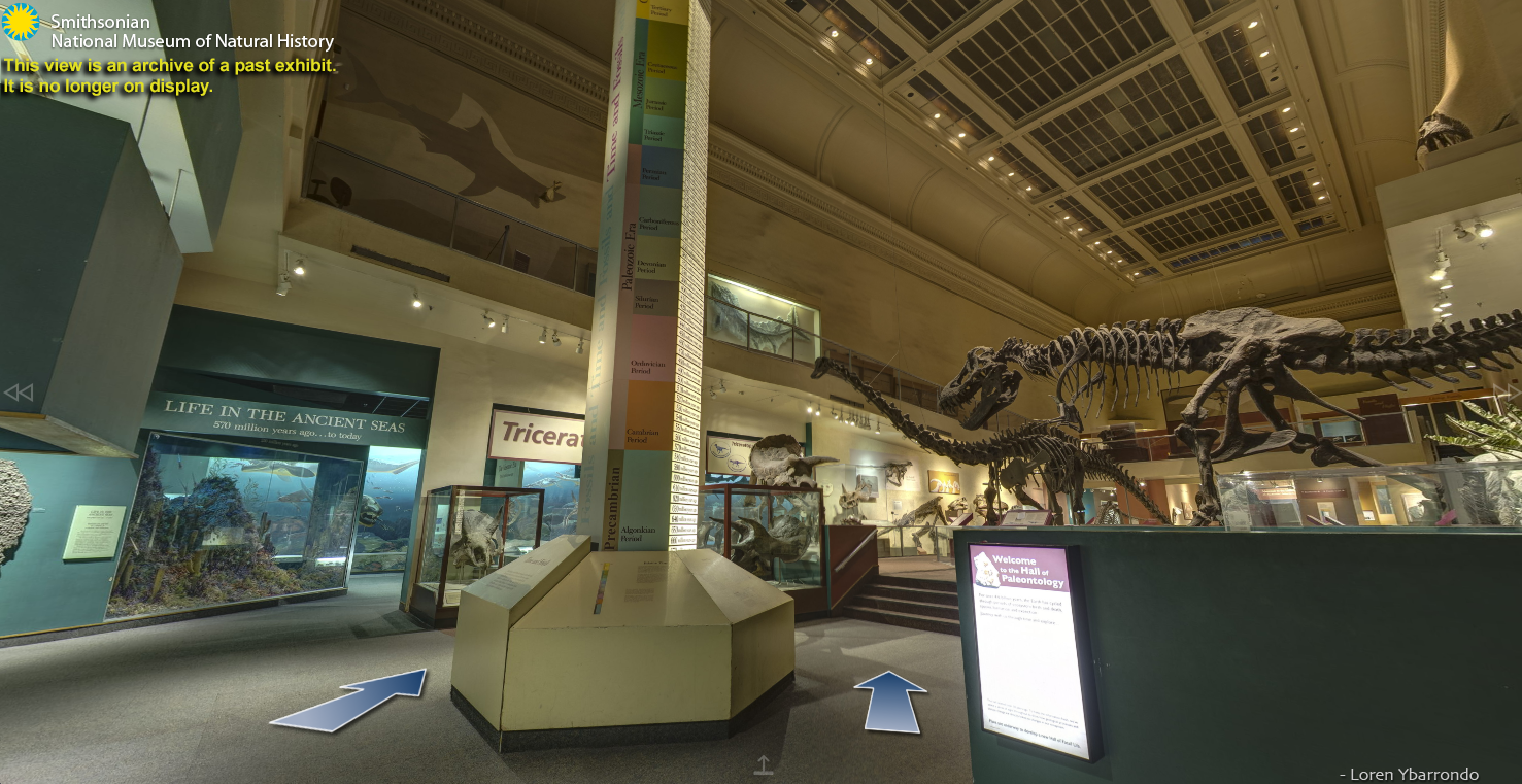 A screenshot from the National Museum of Natural History's Fossil Hall, which closed to the public in 2013 for renovation. This screenshot depics a view from the virtual tour. The image contains a view of the center room of the exhibit, with two arrows on the ground that can be clicked to navigate the exhibit. To the left there is the entrance to a section labeled Life in the Ancient Seas which focuses on prehistoric aquatic life. In the center of the image there is a tower depicting a timeline of different eras of prehistory. The right side of the image includes a sign that says Welcome to the Hall of Paleontology and two large dinosaur fossils- a Tyrannosaurus rex and a Diplodocus.