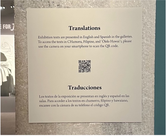 Wall text titled “Translations.” In smaller text below, “Exhibition texts are presented in English and Spanish in the galleries. To access the texts in CHamoru, Filipino, and 'Ōlelo Hawai'I, please use the camera on your smartphone to scan the QR code.” Black and white QR code is directly below. Spanish translation of wall text is under the QR code.